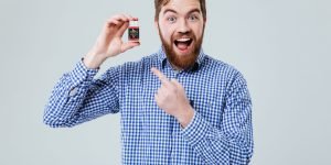 Cheerful excited bearded young man standing and pointing on bottle of pills over white background